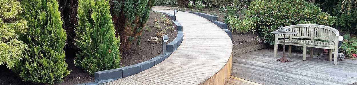 Gebogen houten vlonderpad langs coniferen en een tuinbank